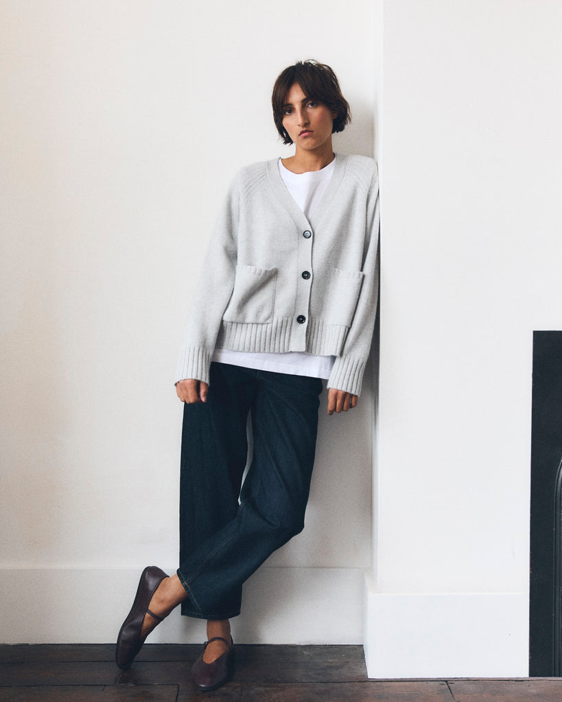 Model with short dark brown hair  leans against white wall, her arms by her side. She is looking to the camera. She is wearing a pale grey v neck cashmere cardigan with pockets and three buttons, a white t-shirt and dark blue jeans. 