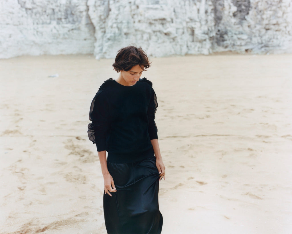 A woman walks on the sand, away from chalky cliffs in the background, her head down and short brown hair blowing slightly in the breeze, she is wearing a black round neck cashmere sweater with transparent black chiffon detail on the shoulders and sleeves which are pushed up on her arms, and she gently handles her black silk skirt between her fingers.