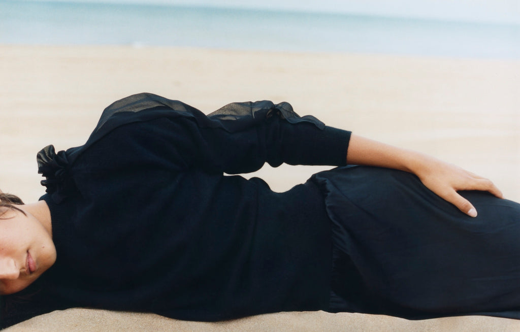 A woman lies on the sand, the grey blue sea hazy in the background, her head rests on her arm and the other hand lies gently on her hip, she is wearing a black round neck cashmere sweater with transparent black chiffon detail on the shoulders and sleeves which are pushed up on her arms, and a black silk skirt.