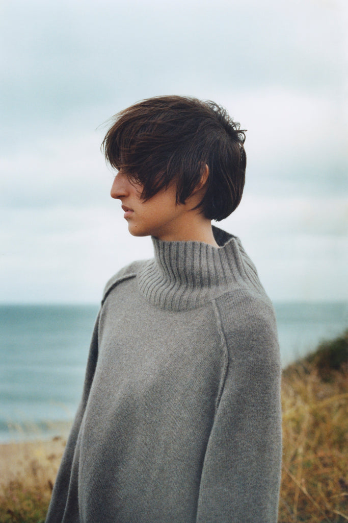 Model stands on cliff in front of sea and cloudy sky. She is looking to the left with short brown hair windswept in face. She is wearing a grey brown oversized high neck sweater with her arms at her sides.