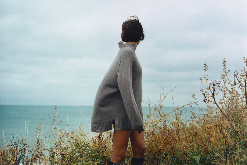 Model stands on cliff in front of sea and cloudy sky in high grasses. She is backwards away from the camera out to sea. She has short brown hair and is wearing a grey brown oversized high neck sweater with her arms at her sides and it is billowing behind her in the wind. Her legs are bare and she is wearing knee high boots that are barely in shot.