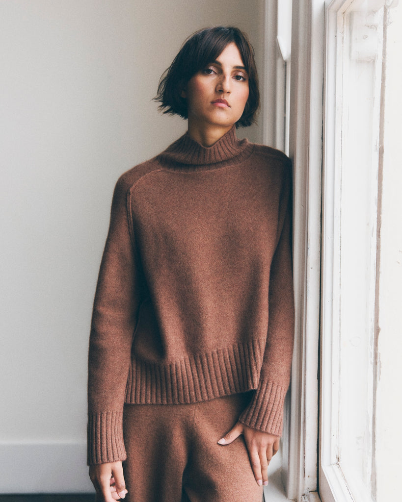 A model stands beside a window in a white room. She has short brown hair and is looking directly at the camera. She is wearing a hazelnut brown cashmere sweater with a relaxed high neck and ribbed hem and cuffs. 