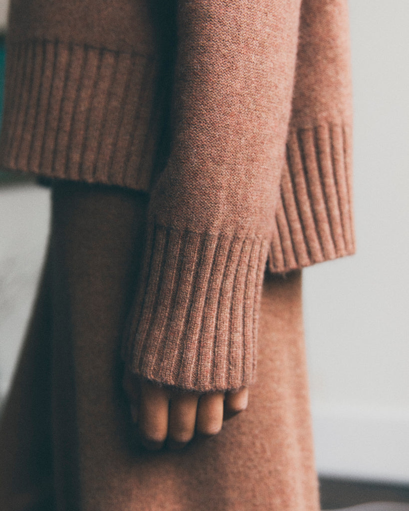 Close up detail of a model standing with her hand by her side. She is wearing a hazelnut brown cashmere sweater with a ribbed hem and cuffs. 