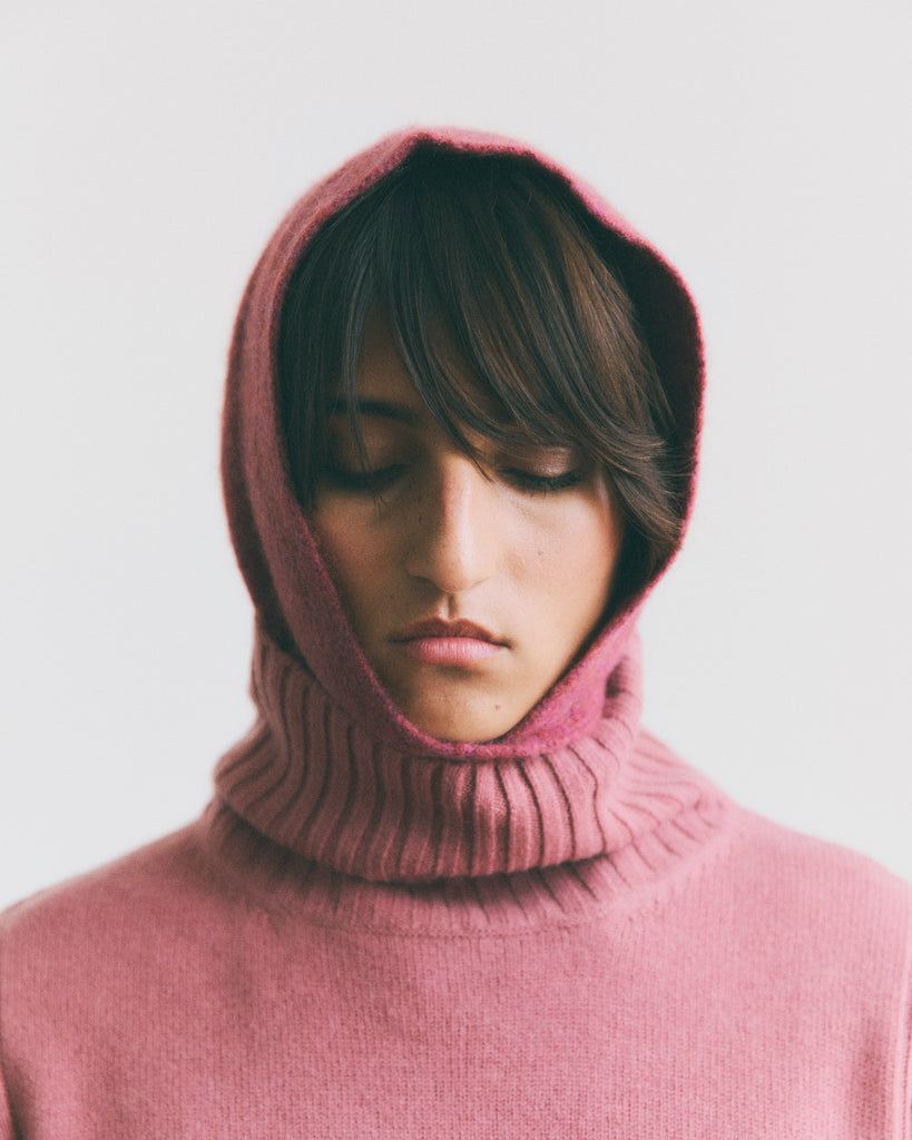 Close up image of model with dark brown floppy fringe hair, her eyes downcast wearing a pink woollen cashmere bonnet snood and a pink high rib neck cashmere sweater 