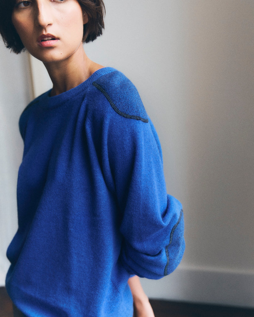 A model with short brown hair standing in a white room looks to the side, she has one arm behind her back and is wearing a blue round neck Sphere One cashmere sweater with patches on the arms and elbows. One sleeve is pushed up. 