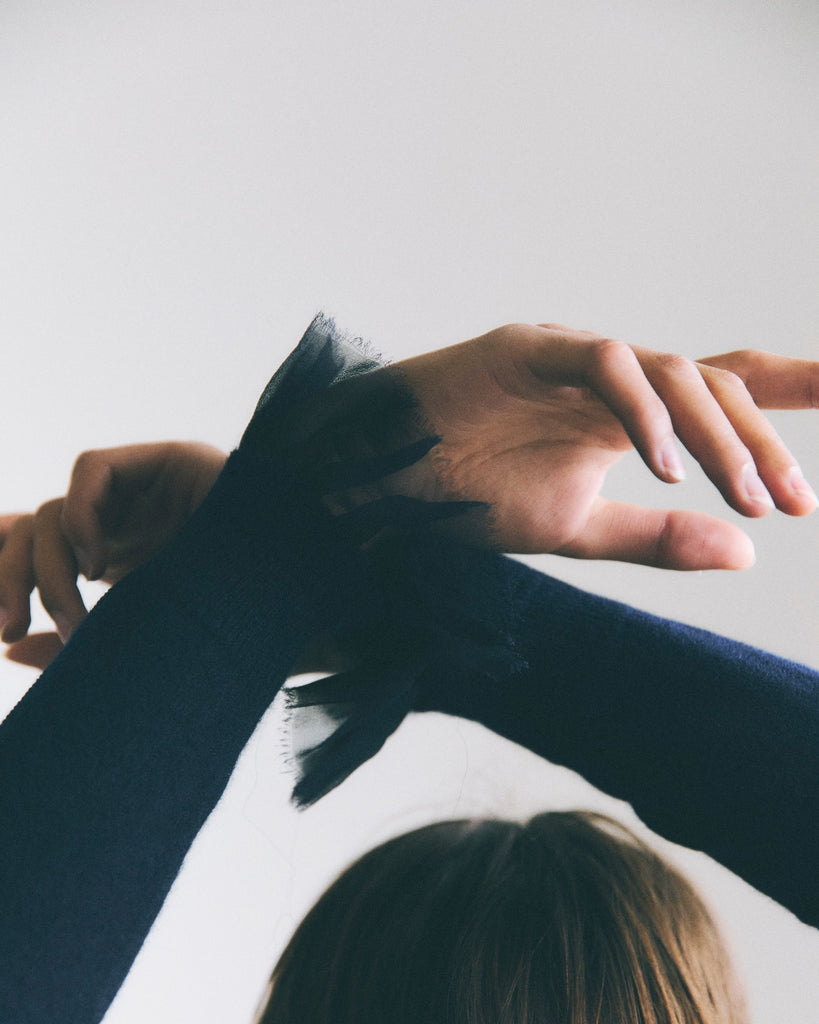 A woman crosses her hands loosely above her head in front of a white background. She has brown hair and is wearing a navy cashmere sweater with navy chiffon cuffs.