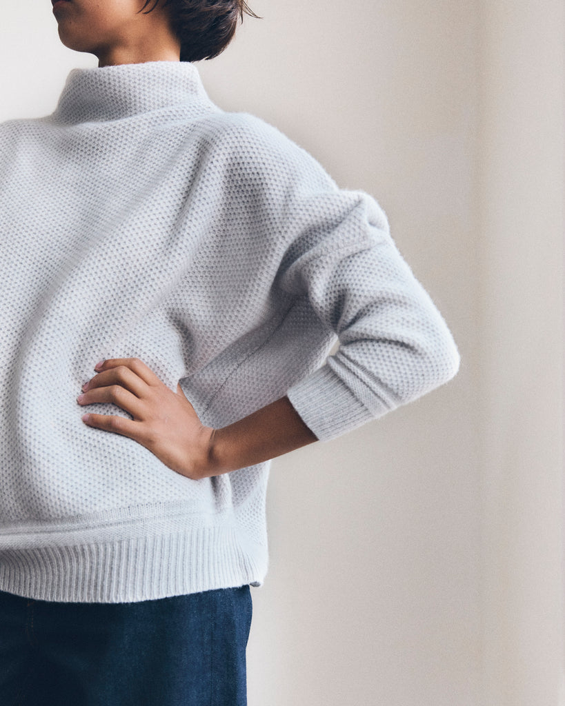 A model stands with her hand on her hip, she has short brown hair and is wearing a light grey honeycomb texture cashmere sweater from Sphere One and dark jeans.  