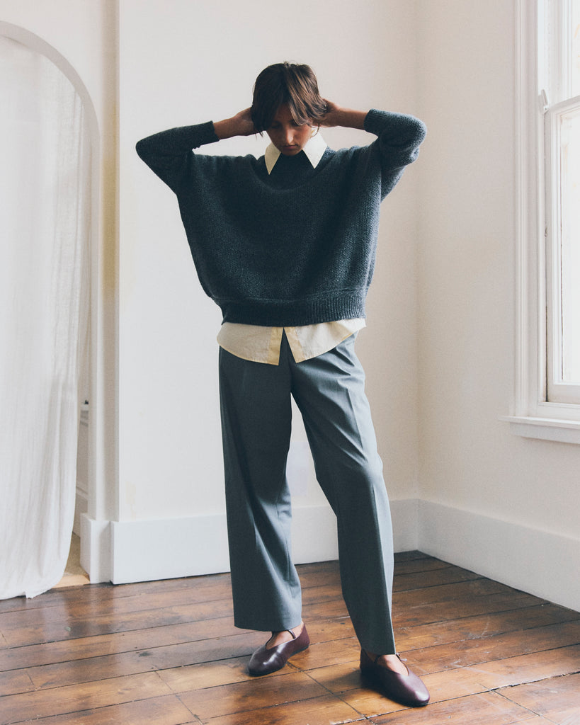 Portrait lookbook shot of model standing beside a window in a room with white walls, a white curtain and wooden floors. She is looking down and has her hands up to her short brown hair. She is wearing a grey Sphere One cashmere sweater with a yellow shirt and grey trousers.