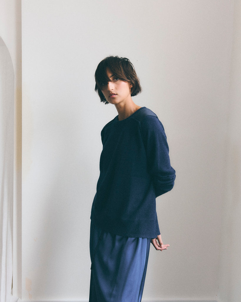 Model with short dark hair in her eyes stands in a white room with her arms loosely behind her back, wearing a navy blue cashmere sweater with navy shoulder patches and a navy silk skirt