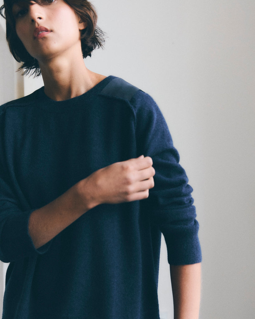 Model with short dark hair looks at the camera, she is pulling at the pushed up sleeve of a navy blue cashmere sweater with grosgrain shoulder patch