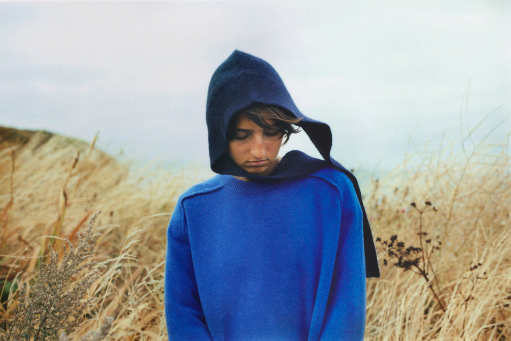 A model wearing a navy bonnet stands in front of sea and cloudy sky among yellow grasses on top of a cliff. She is looking down and wearing a bright blue round neck sweater. Her short brown hair is blowing slightly in the breeze. 