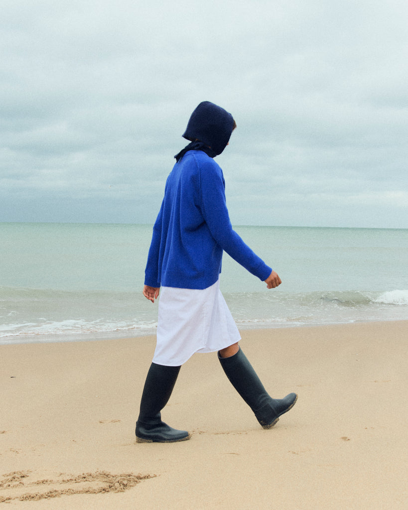 A model wearing a navy bonnet is walking along the beach in profile shot, in front of greyish sea and cloudy sky. She is looking down and wearing a bright blue round neck sweater. and white skirt, with knee high riding boots.   