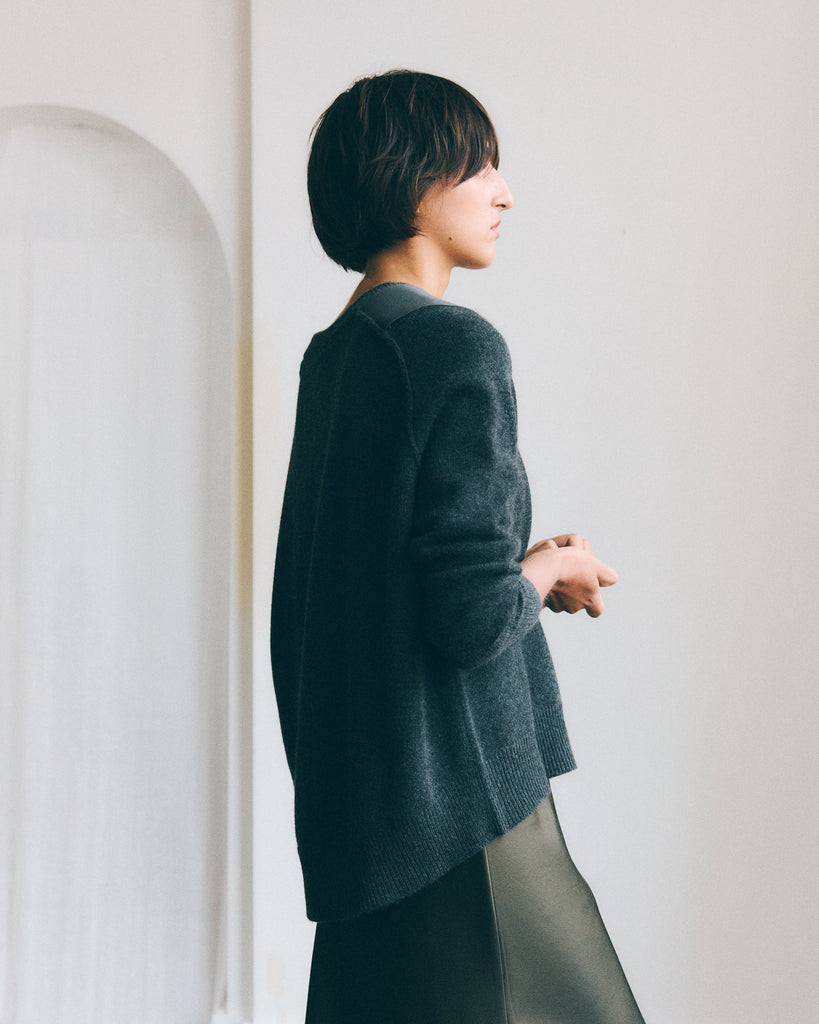 A model stands in profile in a white room with her hands together. She has dark short brown hair and is wearing a Sphere One long green/grey cashmere sweater with a curved back hem and shoulder epaulettes with a green silk skirt. 