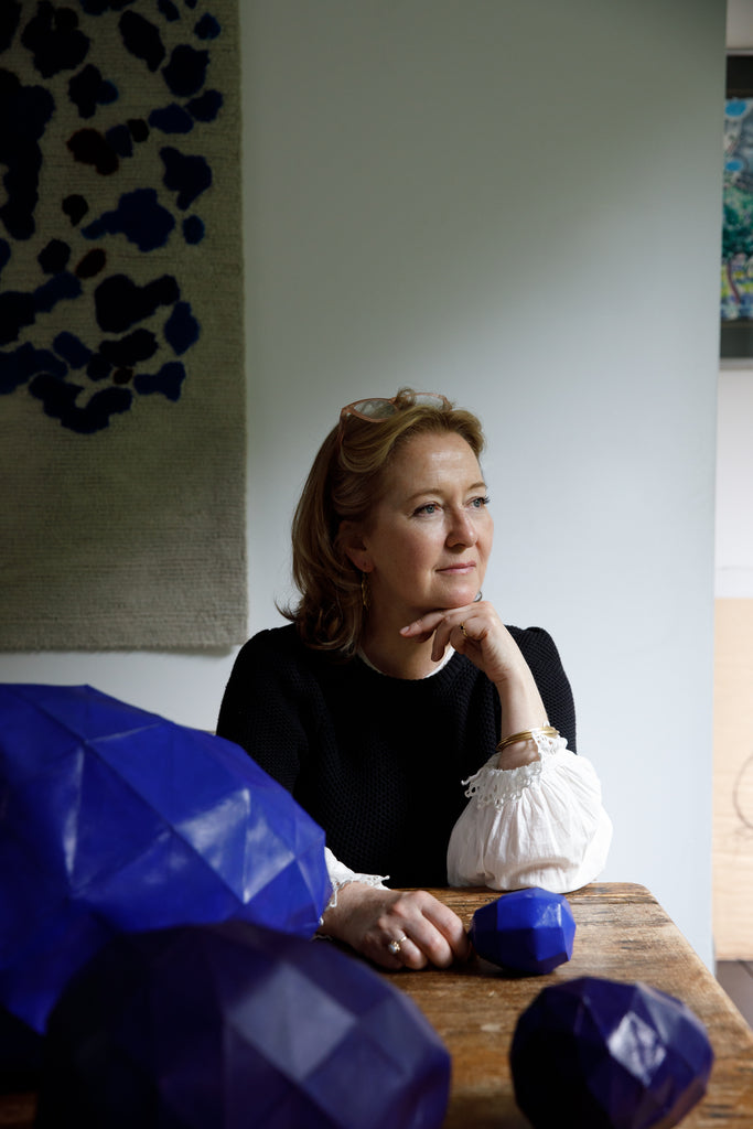 Portrait photo by photographer Doreen Kilfeather of Sphere One designer Lucy Downes. Wearing a dark knitted cashmere sweater, with a white lace cuffed blouse she sits looking to the light, with her elbow on a wooden table, chin on her hand smiling slightly. Her blonde hair is pushed back with by her glasses on her head. A tapestry is seen hanging on a white wall over her shoulder, while in front of her are  selection of blue paper maché geometric eggs.