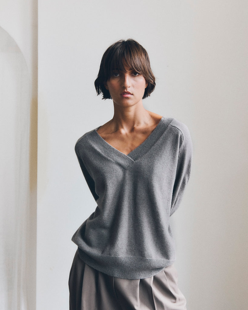 Model stands in front of a white wall, with arms behind her back, looking directly into the camera. She is wearing a light grey deep v neck cashmere sweater. Her neck is bare and she has short brown hair.