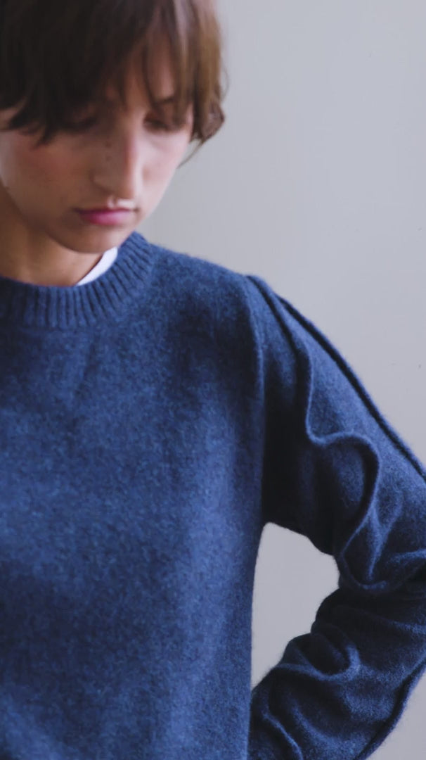 Video of a woman with short brown hair modelling a navy melange round neck cashmere Sphere One sweater with a tube detail snaking up the arm. A white shirt pokes out from the bottom of the sweater over the top of a pair of navy slacks.