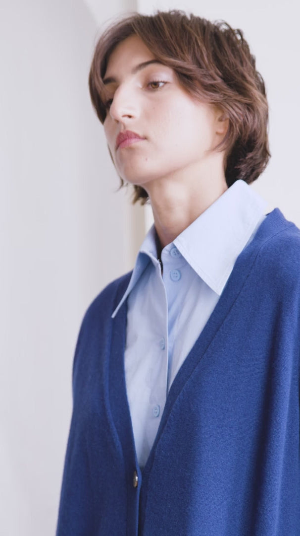 Woman with short dark hair models a blue v neck cashmere cardigan, she is also wearing a light blue shirt with wide lapels 