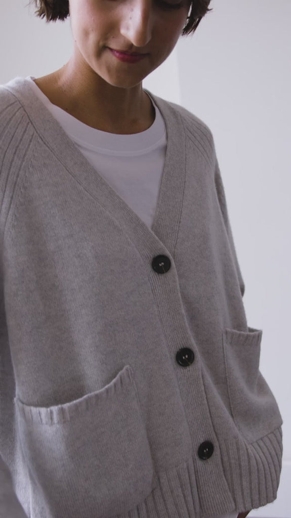 Video of woman with short brown hair modelling a pale grey Sphere One cashmere v neck cardigan with three buttons and front pockets 